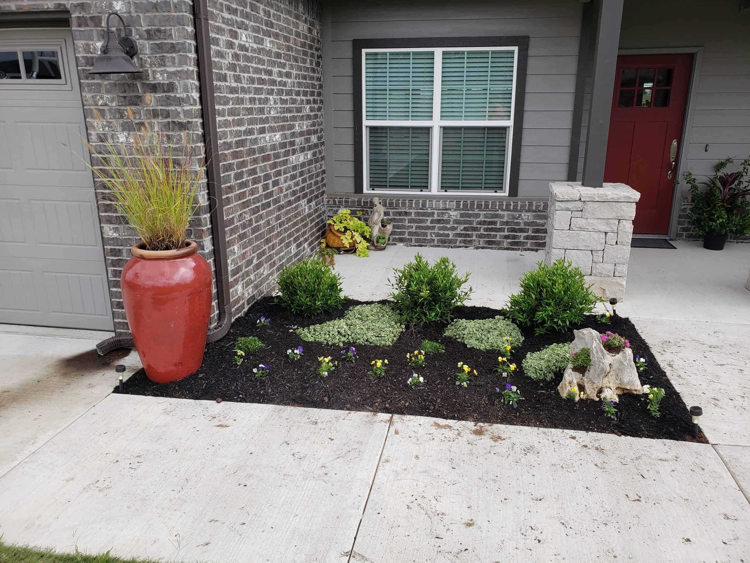 a front patio with a red vase and plants inside a lined landscape bed covered in black mulch